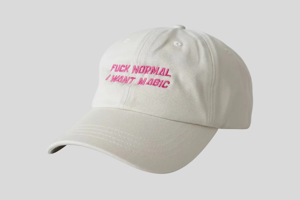 Beige cap with pink text 'F*CK NORMAL I WANT MAGIC' on a carpeted floor.